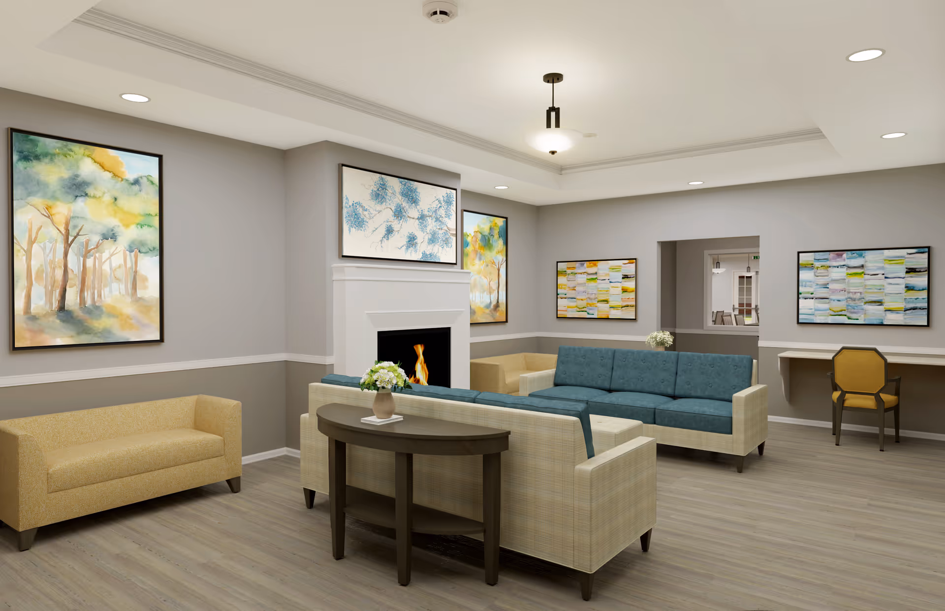 A modern senior living facility common area with a beige and teal sectional sofa arranged around a white fireplace with a fire burning. The room has light gray walls with white trim, wood flooring, and several colorful abstract paintings on the walls. There is a small wooden console table with a vase of flowers behind the sofa, and a yellow chair at a built-in desk area on the right side of the room. Recessed lighting and a ceiling light fixture illuminate the space.