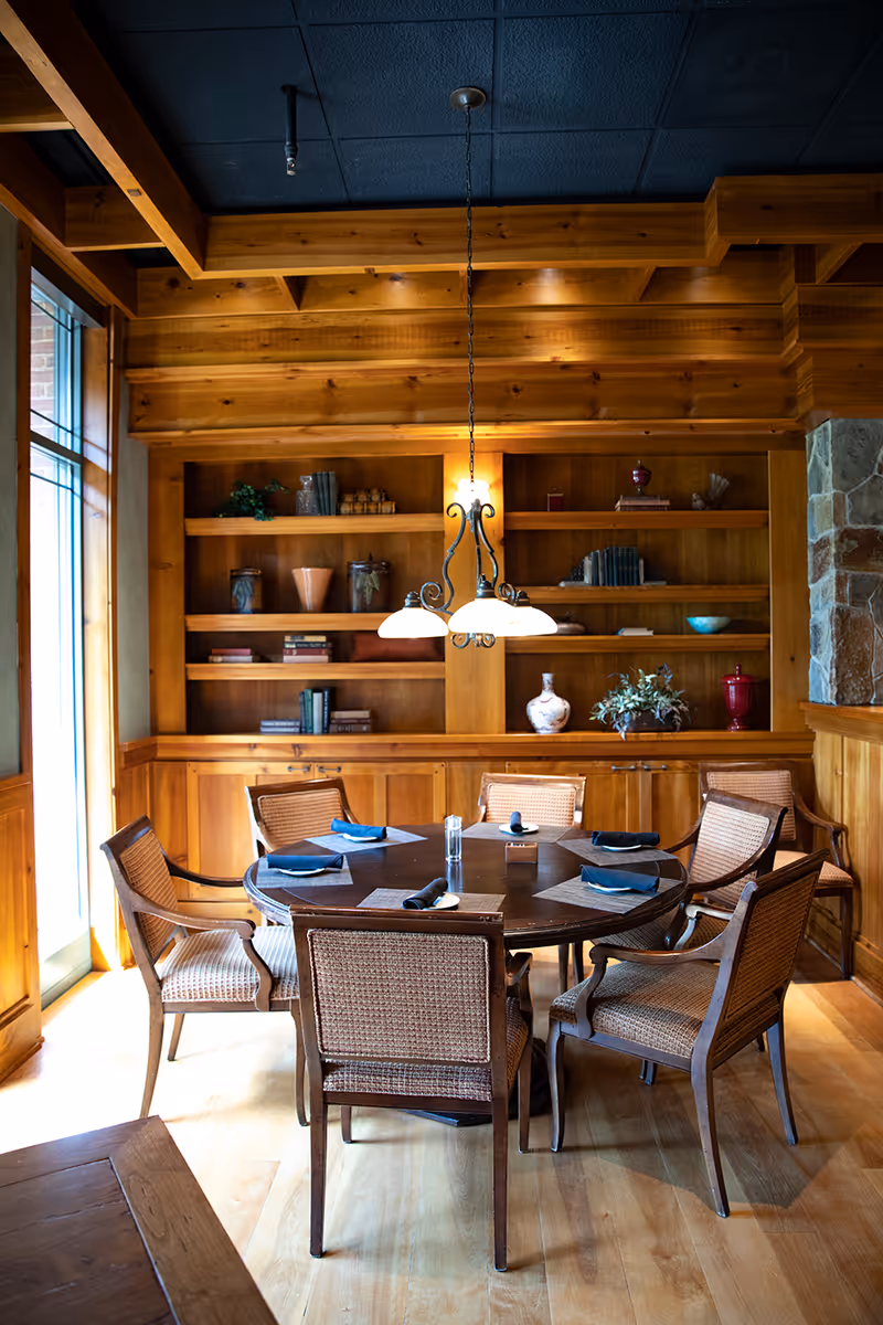 A cozy dining area with a round wooden table set with six chairs. The table has place settings with napkins and a small centerpiece. Behind the table is a wooden built-in bookshelf with various decorative items and books. A hanging light fixture with three lamps illuminates the table. The room features wood paneling and a stone accent on the right side.