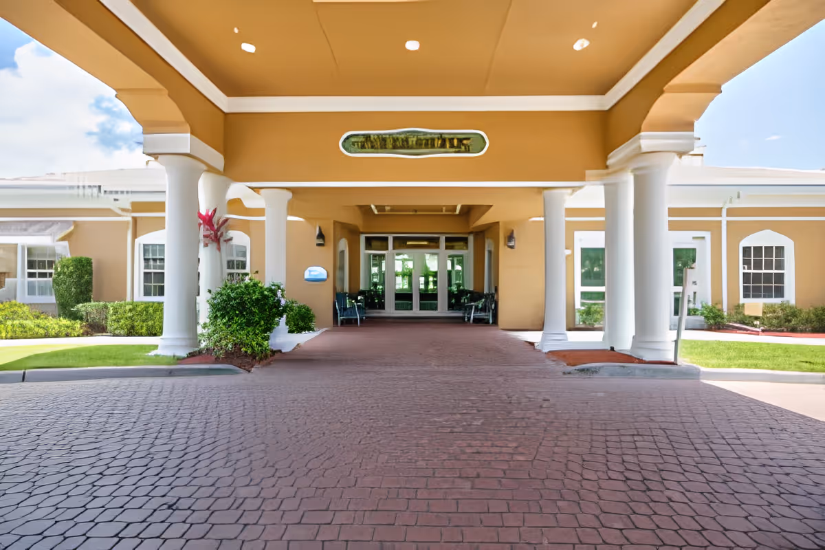 Covered entrance area of a building with white columns and a brick-paved driveway leading to glass double doors. The building exterior is beige with white trim, and there are green bushes and grass on either side of the entrance.