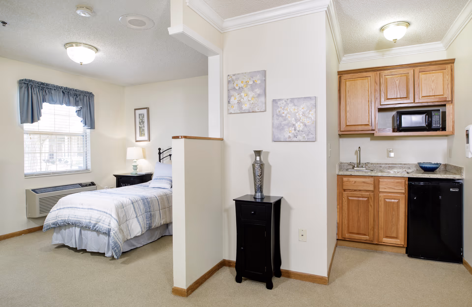 A small assisted living studio apartment featuring a single bed with a plaid comforter near a window with blue curtains. Next to the bed is a nightstand with a lamp. A half wall separates the sleeping area from a kitchenette with wooden cabinets, a microwave, a small sink, and a mini refrigerator. A black side table with a decorative vase and two floral paintings are on the wall dividing the two spaces.