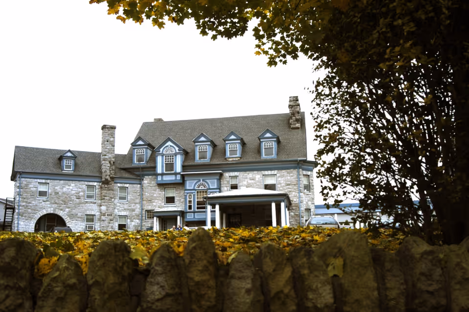 Stone building with multiple windows and a steep roof, partially obscured by a stone fence and autumn leaves in the foreground, with trees framing the right side of the image.