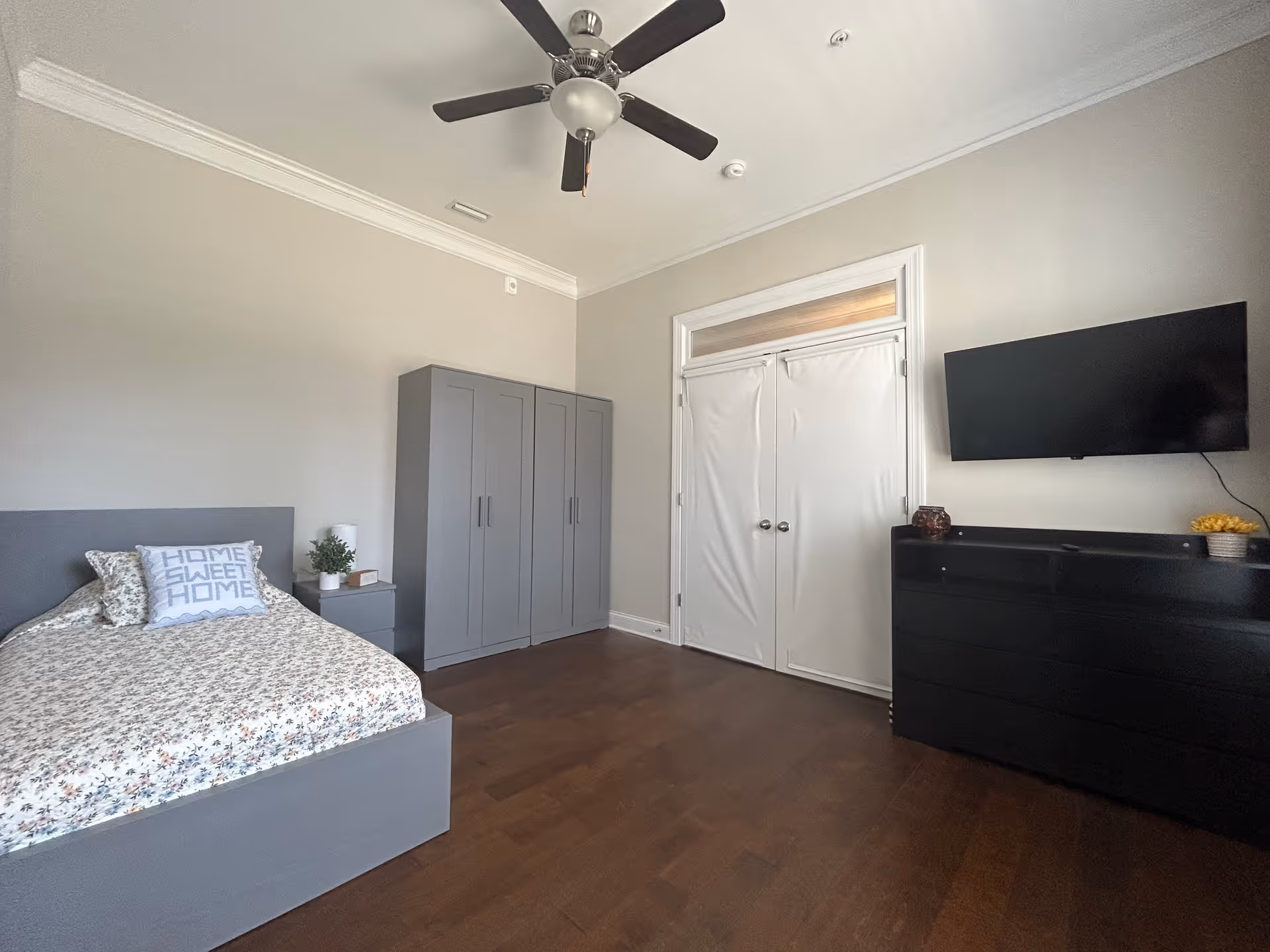 A bedroom with a gray bed frame and floral bedding, a pillow with the text 'HOME SWEET HOME', a gray wardrobe, a nightstand with a small plant and lamp, a ceiling fan, double white doors, a black dresser with decorative items, and a wall-mounted flat-screen TV.
