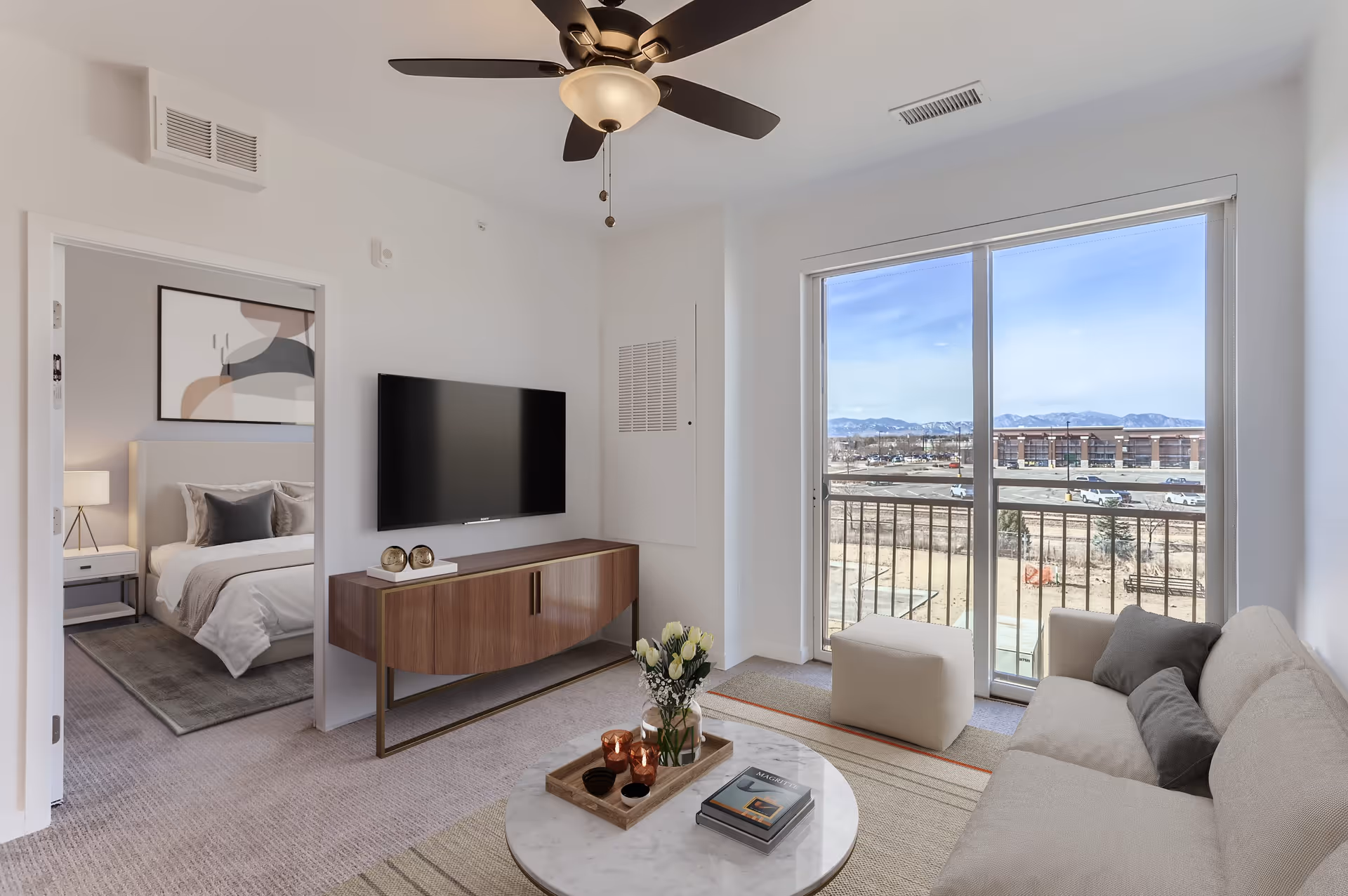 A bright living room with a beige sofa, a round marble coffee table with decorative items, a wooden TV console with a flat-screen TV mounted above it, and a ceiling fan with lights. There is a large sliding glass door leading to a balcony with a view of a parking lot and mountains in the distance. An open doorway reveals a bedroom with a bed, nightstand, lamp, and abstract wall art.