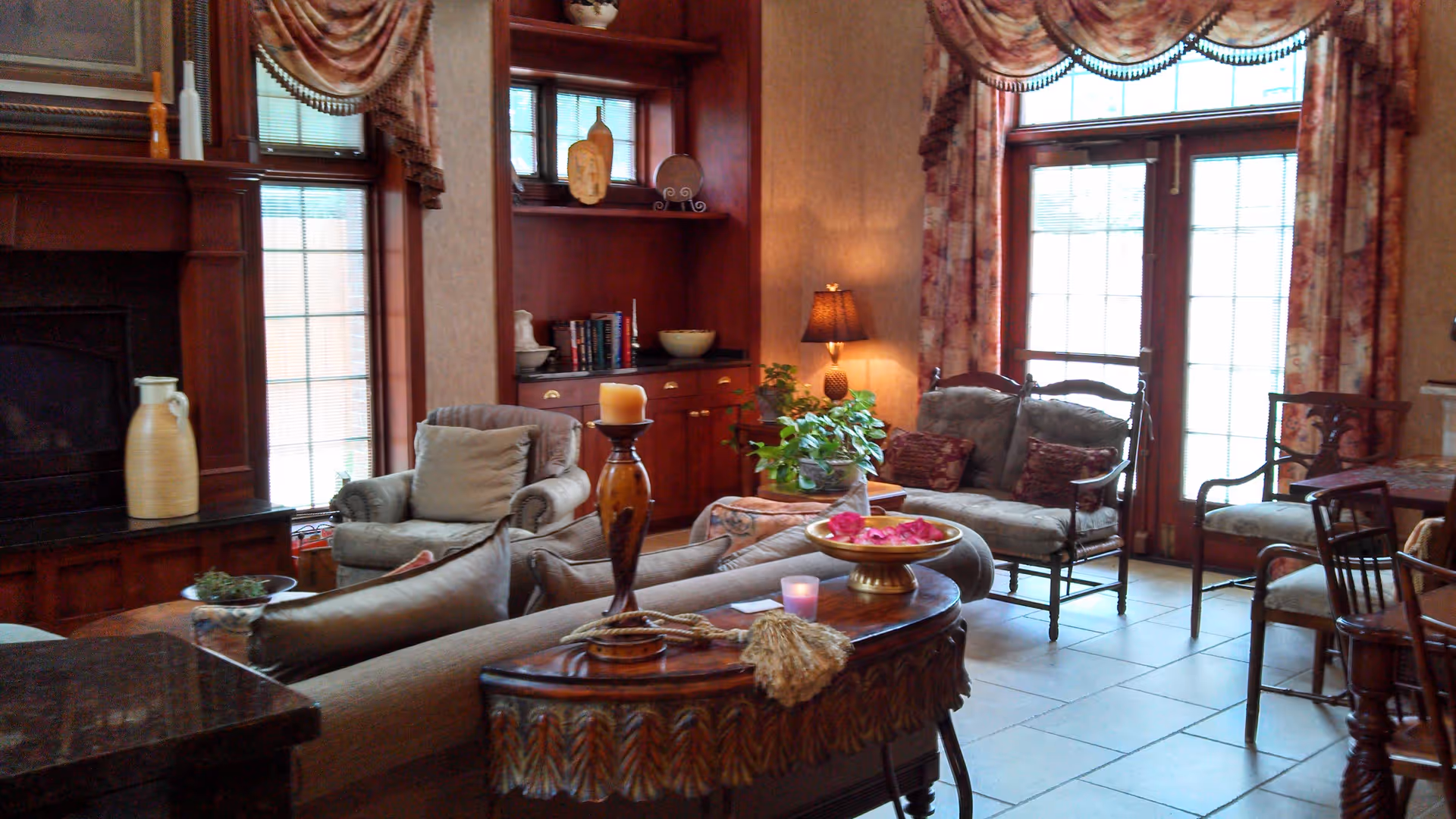 A cozy living room with a fireplace, upholstered armchairs, a wooden coffee table with decorative items, potted plants, and large windows with floral curtains allowing natural light to enter.