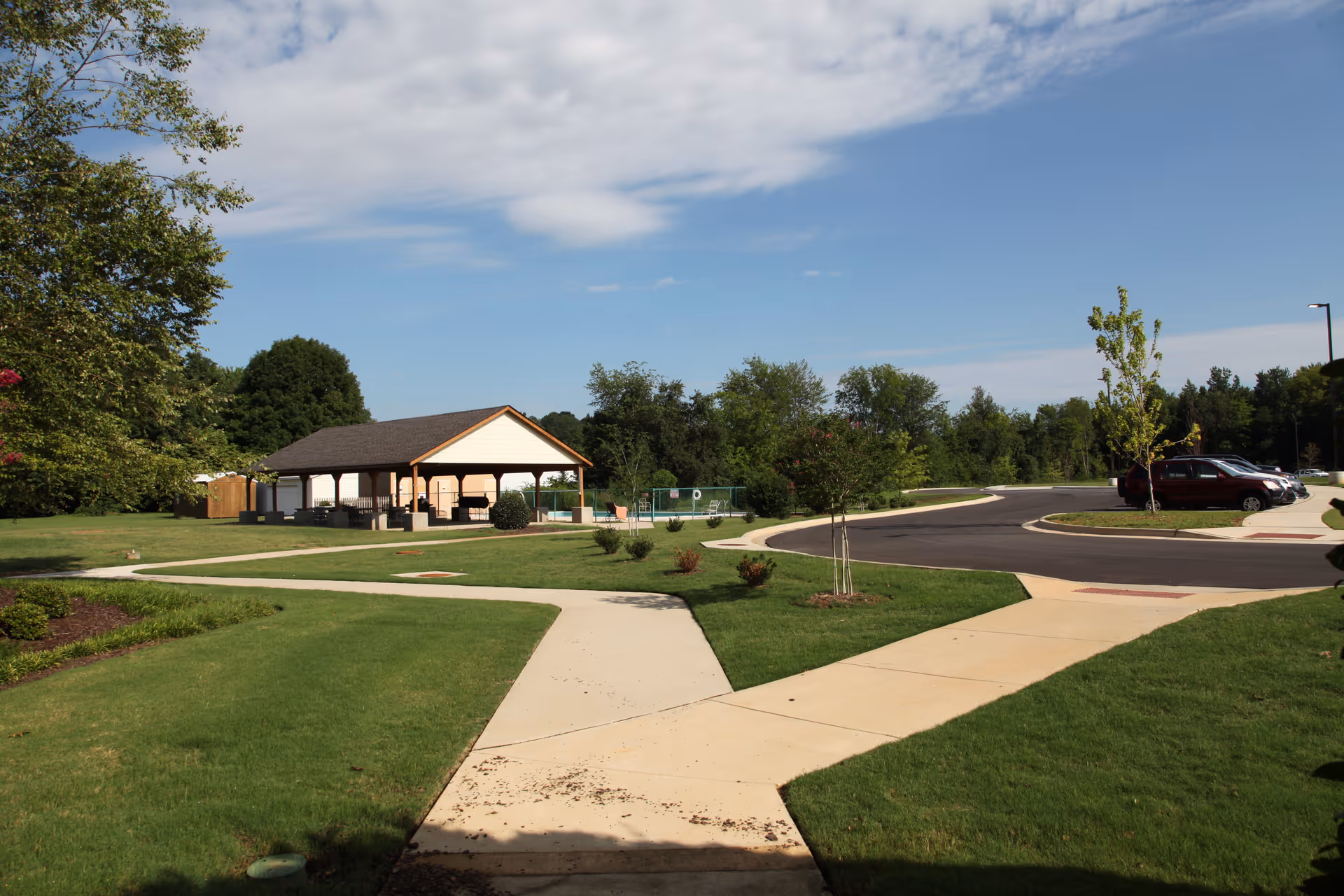 Outdoor view of a senior living facility with a paved walkway, green lawns, small trees, a covered pavilion with seating, a fenced pool area, and a parking lot with several cars under a partly cloudy sky.
