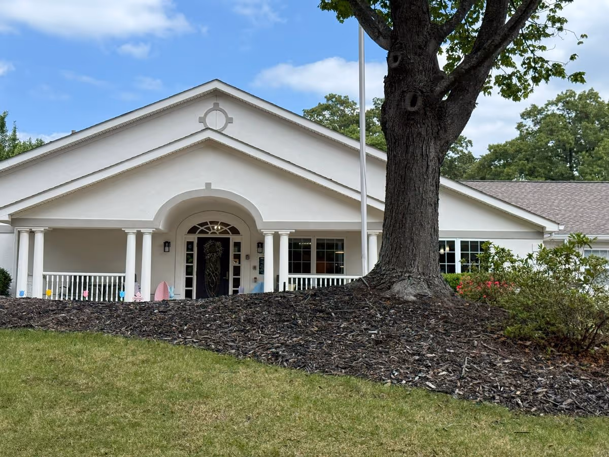 White single-story building with a columned front porch, flagpole, and a large tree in the foreground.