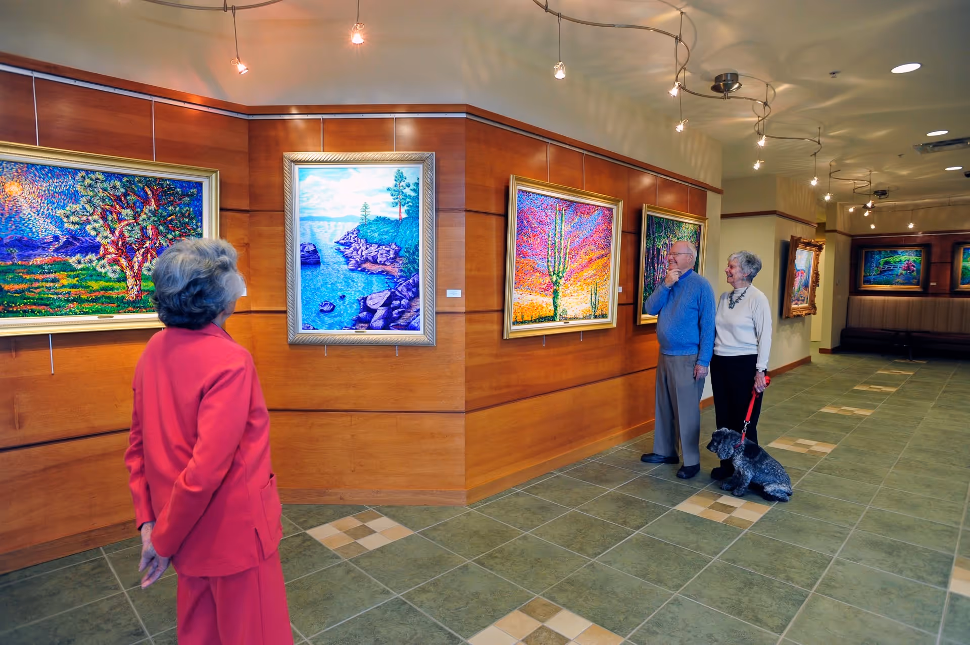 Three older adults, one with a small dog, view colorful framed paintings displayed on a wood-paneled gallery wall in a tiled hallway.
