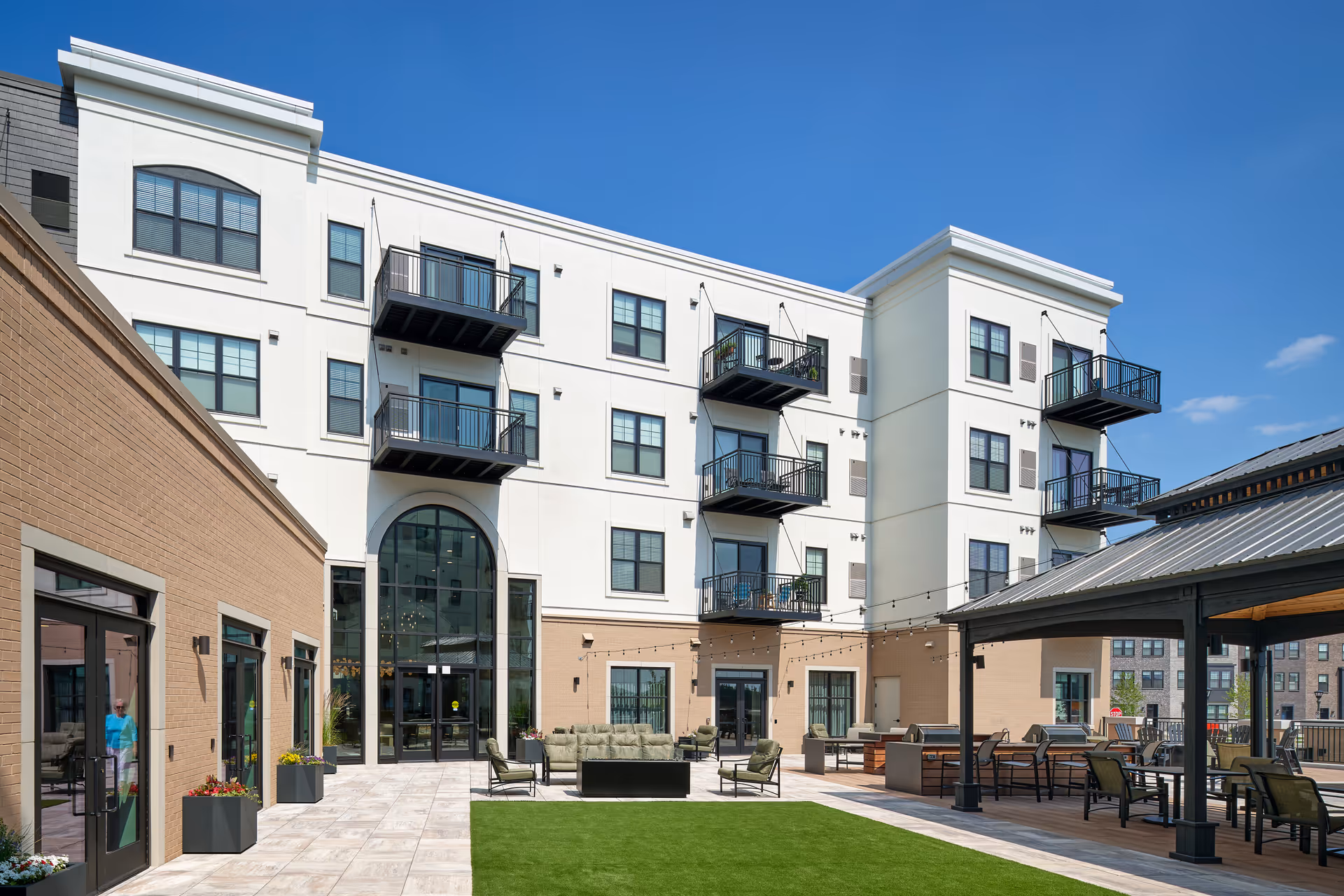 Outdoor courtyard area of a senior living facility named Marvella, featuring a green lawn, patio seating with chairs and sofas, string lights overhead, and a multi-story building with balconies and large windows under a clear blue sky.