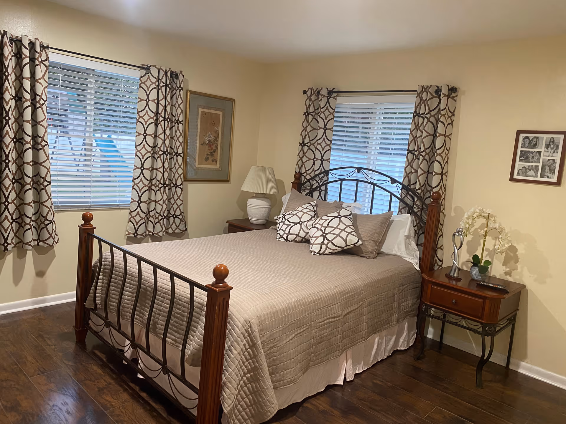A neatly made bed with a metal and wood frame in a bedroom. The bed has a beige quilt and matching pillows with a geometric pattern. There are two windows with blinds and patterned curtains on adjacent walls. A wooden nightstand with a white lamp and a small decorative plant is beside the bed. The room has wooden flooring and light-colored walls with framed artwork and photos.