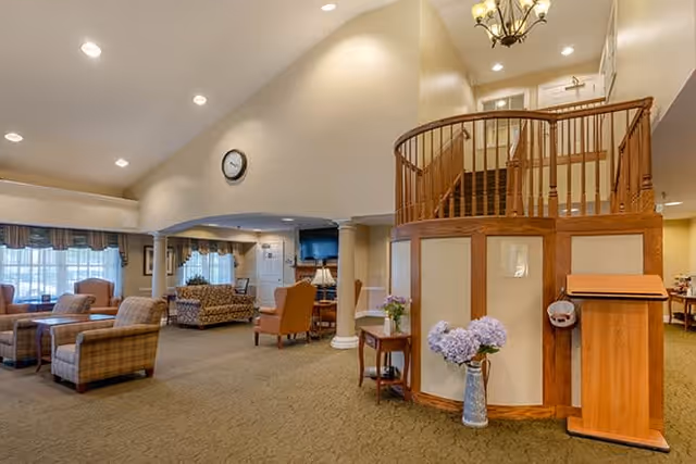 Spacious senior living facility common area with comfortable upholstered chairs and sofas arranged around coffee tables. The room features high ceilings with recessed lighting, a wall clock, large windows with curtains, and a wooden staircase with a curved railing leading to an upper floor. There is a small table with a vase of flowers and a wooden podium near the staircase.