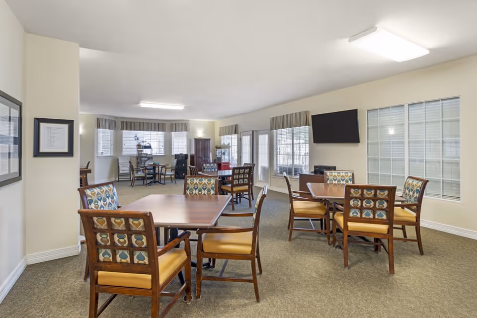 Well-lit common dining/activity room with several wooden tables and patterned chairs, large windows and a wall-mounted TV.