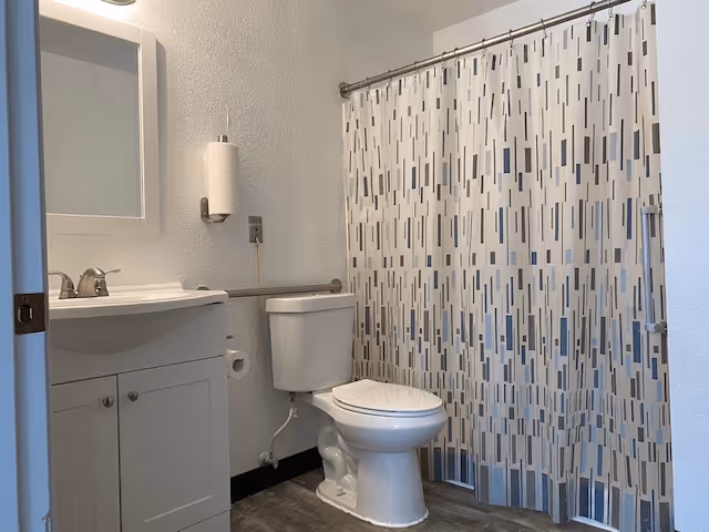 A clean bathroom featuring a white toilet, a white vanity with a sink and faucet, a wall-mounted mirror above the sink, a paper towel holder with a roll of paper towels, and a shower area with a patterned shower curtain in shades of white, gray, and blue.