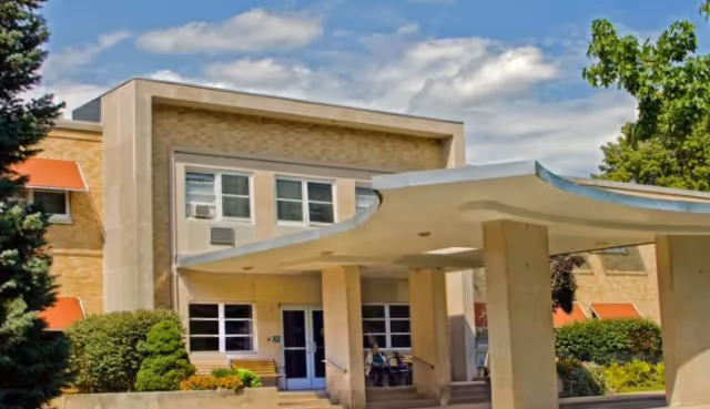 Entrance to a beige-brick senior care facility with a curved white canopy, columns, and landscaping.