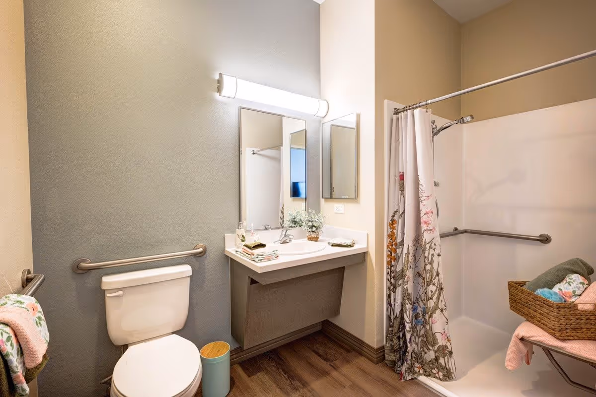 A clean and accessible bathroom featuring a toilet with grab bars, a sink with a large mirror and overhead light, a shower with a floral curtain and grab bars, and a basket with towels on a small bench.