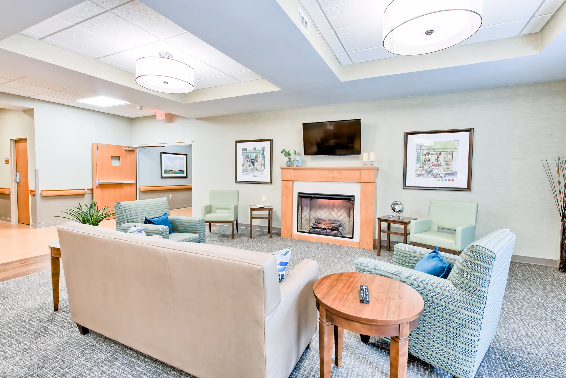A cozy senior living facility lounge area with a beige sofa, two striped armchairs with blue pillows, and two light green armchairs. The room features a wooden fireplace with a TV mounted above it, two framed pictures on the wall, and two round ceiling lights. There are small wooden side tables with decorative items and a remote control on one table. The floor is carpeted and the walls are light-colored.