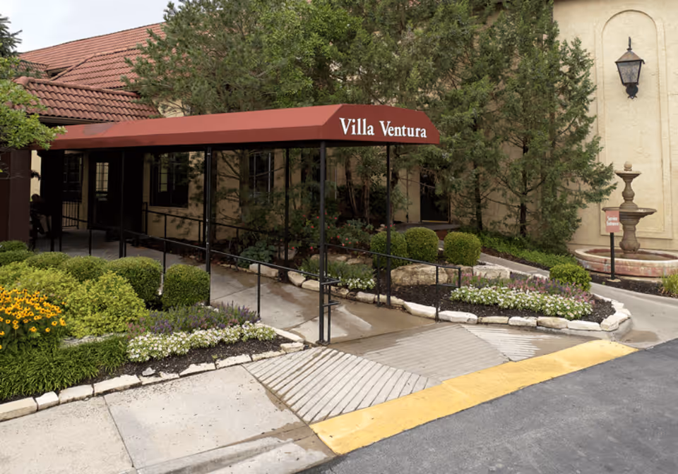 Entrance to Senior Star at Villa Ventura with a red canopy labeled 'Villa Ventura' over a walkway surrounded by landscaped bushes, flowers, and trees. There is a beige building with a tiled roof and a decorative fountain near the entrance.