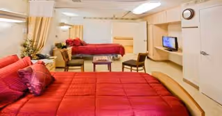 Two red bedspreads cover patient beds in a communal senior living bedroom with chairs and TV.