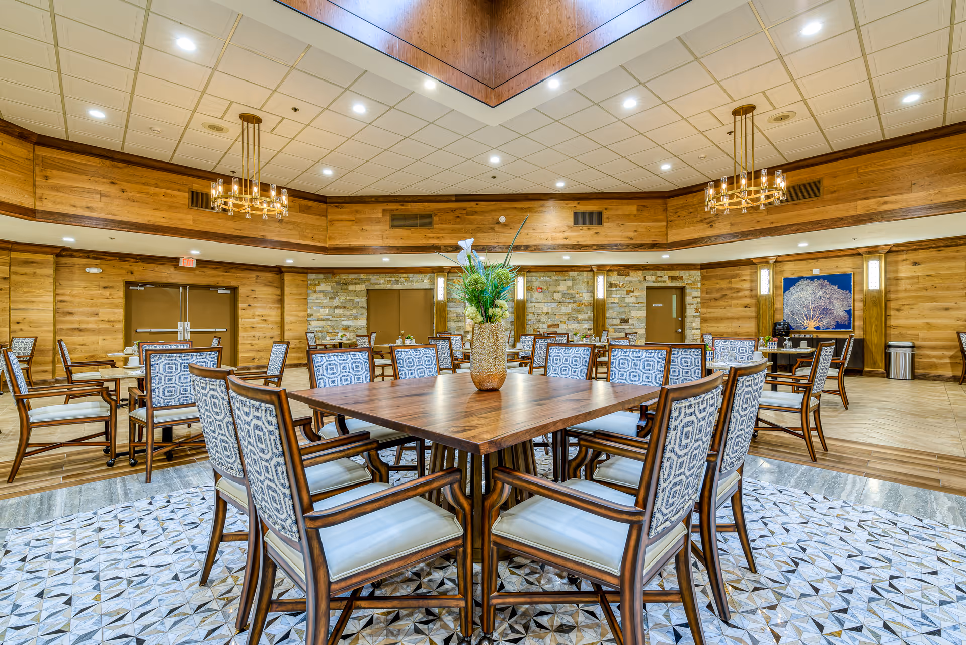 A spacious dining room with wooden walls and ceiling, featuring multiple tables surrounded by cushioned chairs with patterned backs. A large square table with a floral centerpiece is in the foreground. The room is well-lit with recessed lighting and two chandeliers, and has a patterned tile floor area beneath the main table.