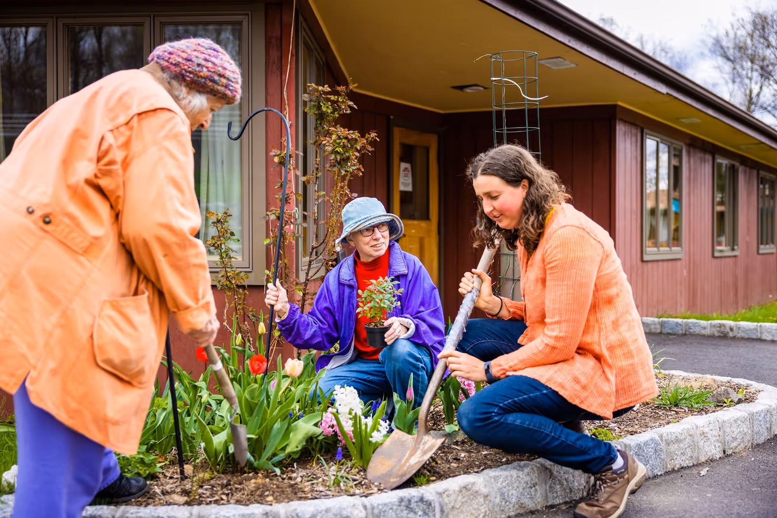 Three women gardening together outside a building with red siding. One woman is holding a small potted plant, another is digging with a shovel, and the third is working with a garden tool. They are surrounded by blooming flowers and greenery.
