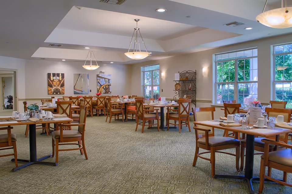 Spacious assisted-living dining room with multiple set tables, wooden chairs, large windows, and pendant lights.