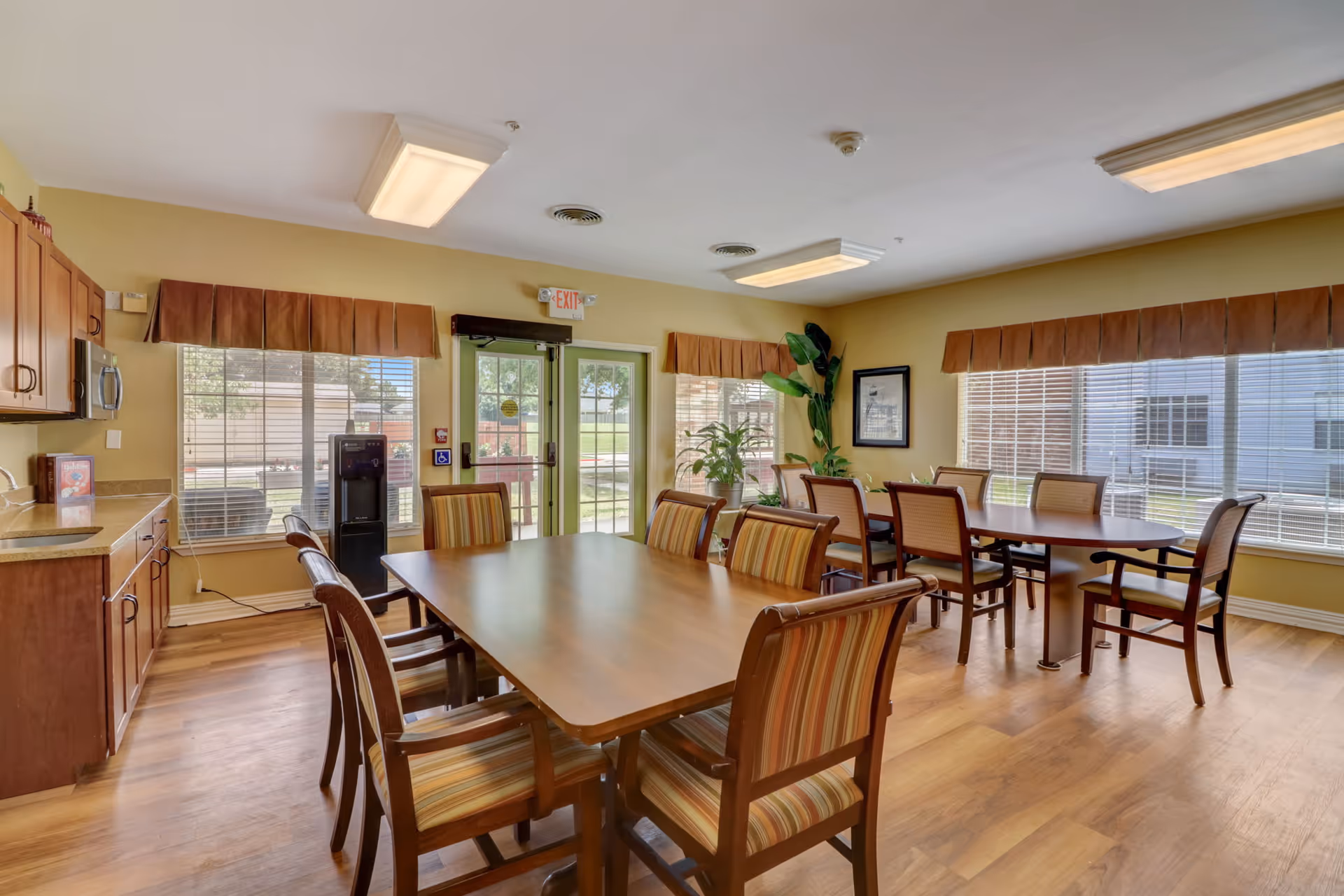 A bright dining room with two wooden tables surrounded by striped cushioned chairs. The room has large windows with blinds and valances, a wooden floor, and a small kitchenette area with cabinets and a countertop on the left. There are plants near the windows and an exit door with a green frame in the background.