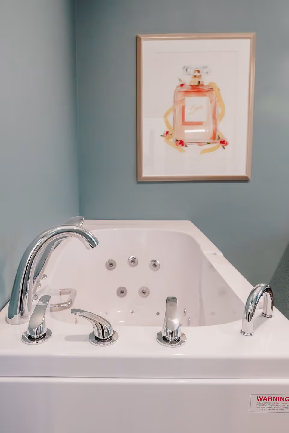 A white whirlpool bathtub with chrome faucets and controls set against a teal wall. Above the bathtub, there is a framed picture of a perfume bottle labeled 'Love' with decorative flowers around it.