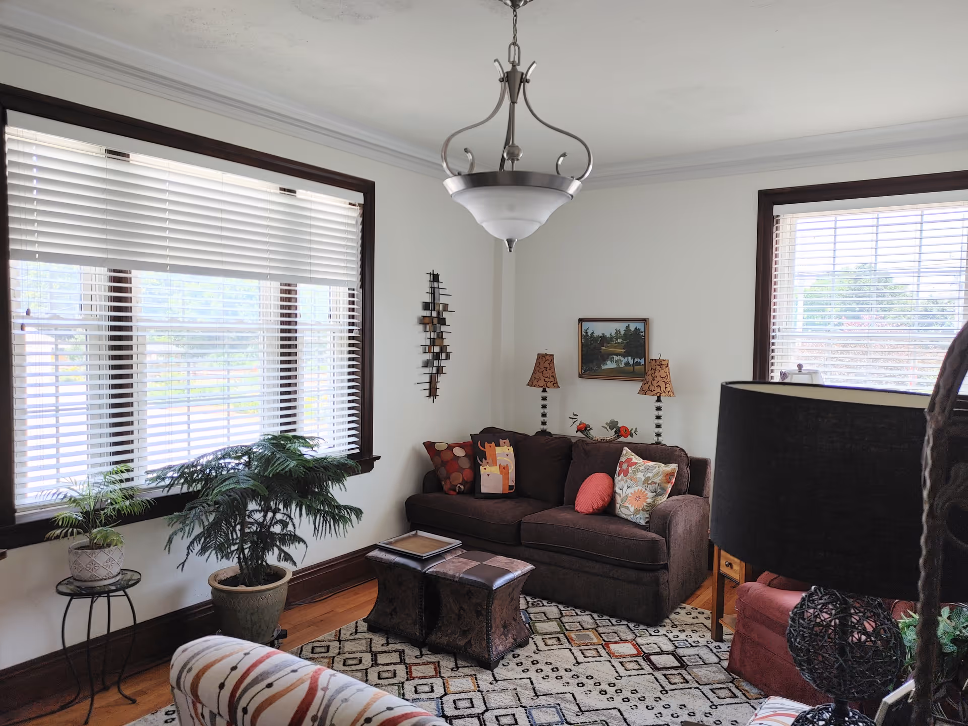 A cozy living room with a dark brown sofa adorned with colorful pillows, two small ottomans, a patterned area rug, and two large windows with white blinds. There are two table lamps with patterned shades on either side of the sofa, a framed landscape painting on the wall, a hanging ceiling light fixture, and several potted plants near the windows.