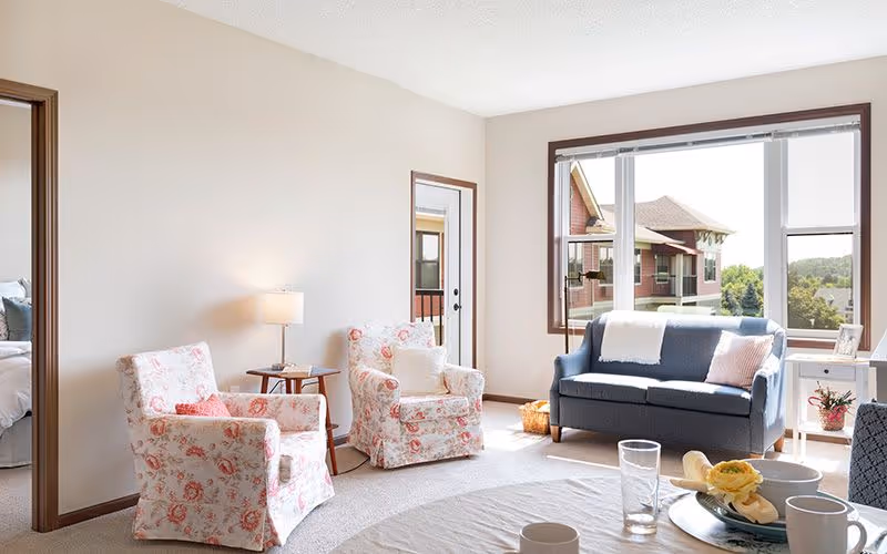 A bright living room with large windows showing an outdoor view. The room features a blue sofa with pillows, two floral armchairs, a round table with cups and a flower centerpiece, a side table with a lamp, and a door leading to another room with a bed visible.