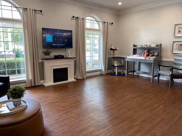A spacious living room with wooden flooring, two large arched windows with beige curtains, a wall-mounted flat screen TV above a white electric fireplace, a small cat tree with cats, a changing table with baby dolls and decorations, and a chair with striped upholstery.