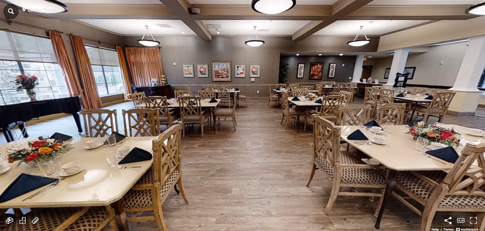 A spacious dining room with multiple tables and chairs arranged neatly. Each table is set with black napkins, white cups, saucers, and glasses. There are floral centerpieces on some tables. Large windows with orange curtains allow natural light to fill the room. The floor is wooden, and the walls are decorated with framed artwork. A black grand piano is visible near the windows.