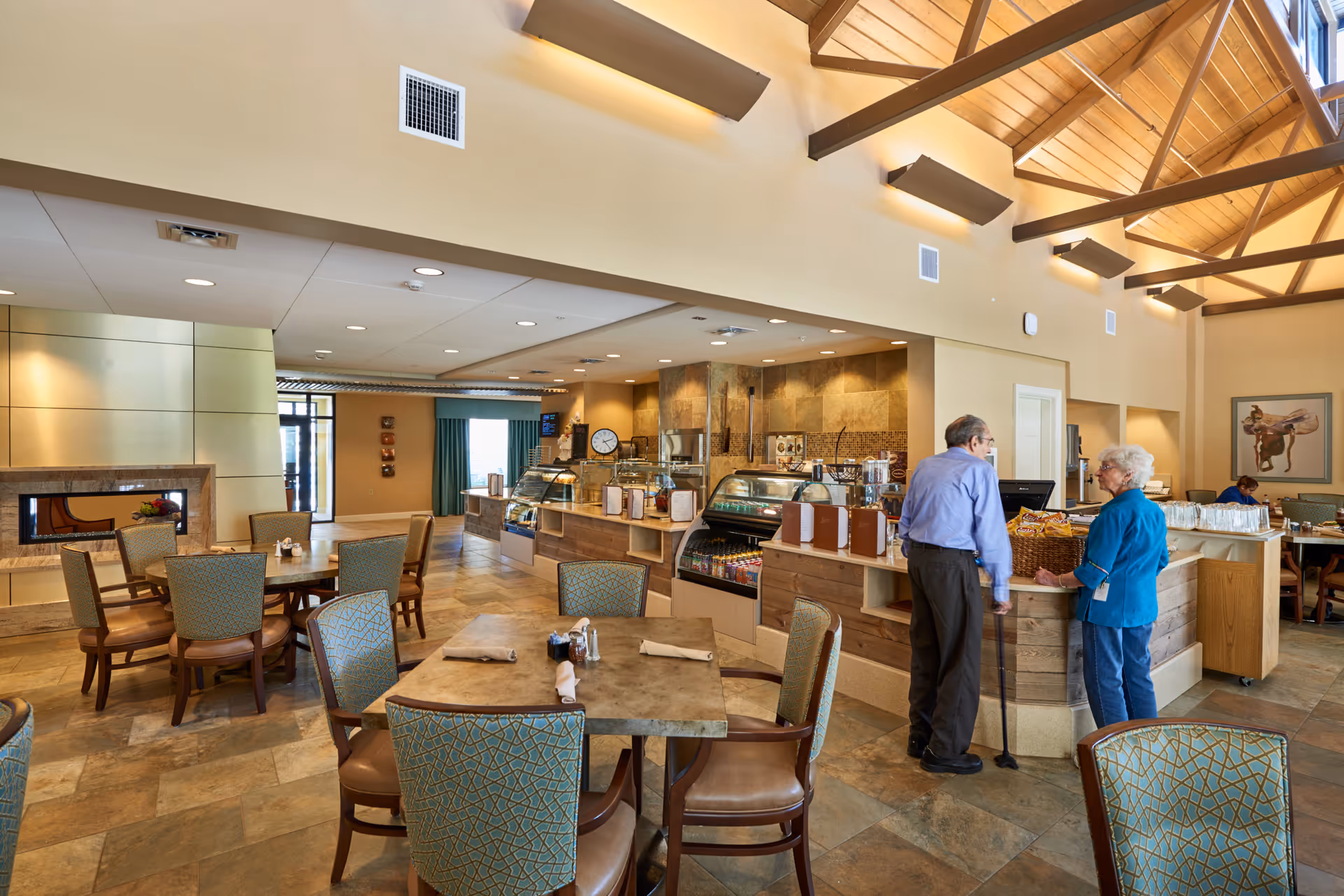 A spacious dining area in an assisted living facility with several tables and chairs. Two elderly people are standing and talking near a counter that has food and drink displays. The room has a high ceiling with wooden beams and warm lighting.