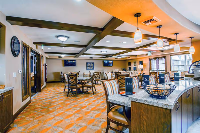 A spacious dining room with multiple tables and chairs, featuring a patterned carpet in orange and beige tones. The ceiling has wooden beams and hanging pendant lights. There are two wall-mounted televisions and decorative artwork on the walls. A countertop with chairs and menus is visible in the foreground.