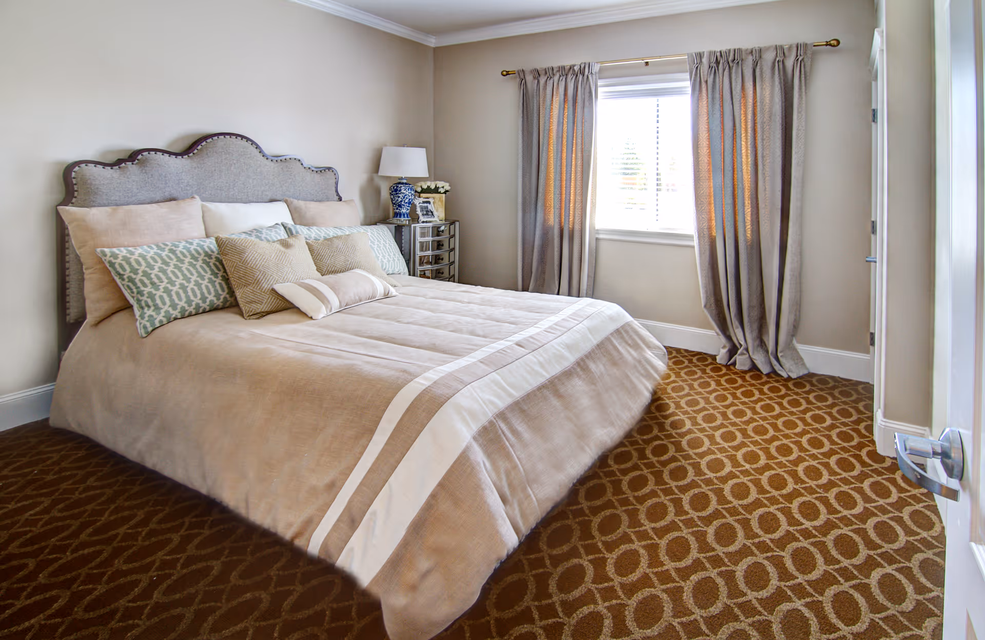 A neatly made bed with multiple pillows in a bedroom featuring beige walls, a window with curtains, a bedside table with a lamp and decorative items, and a patterned carpet.