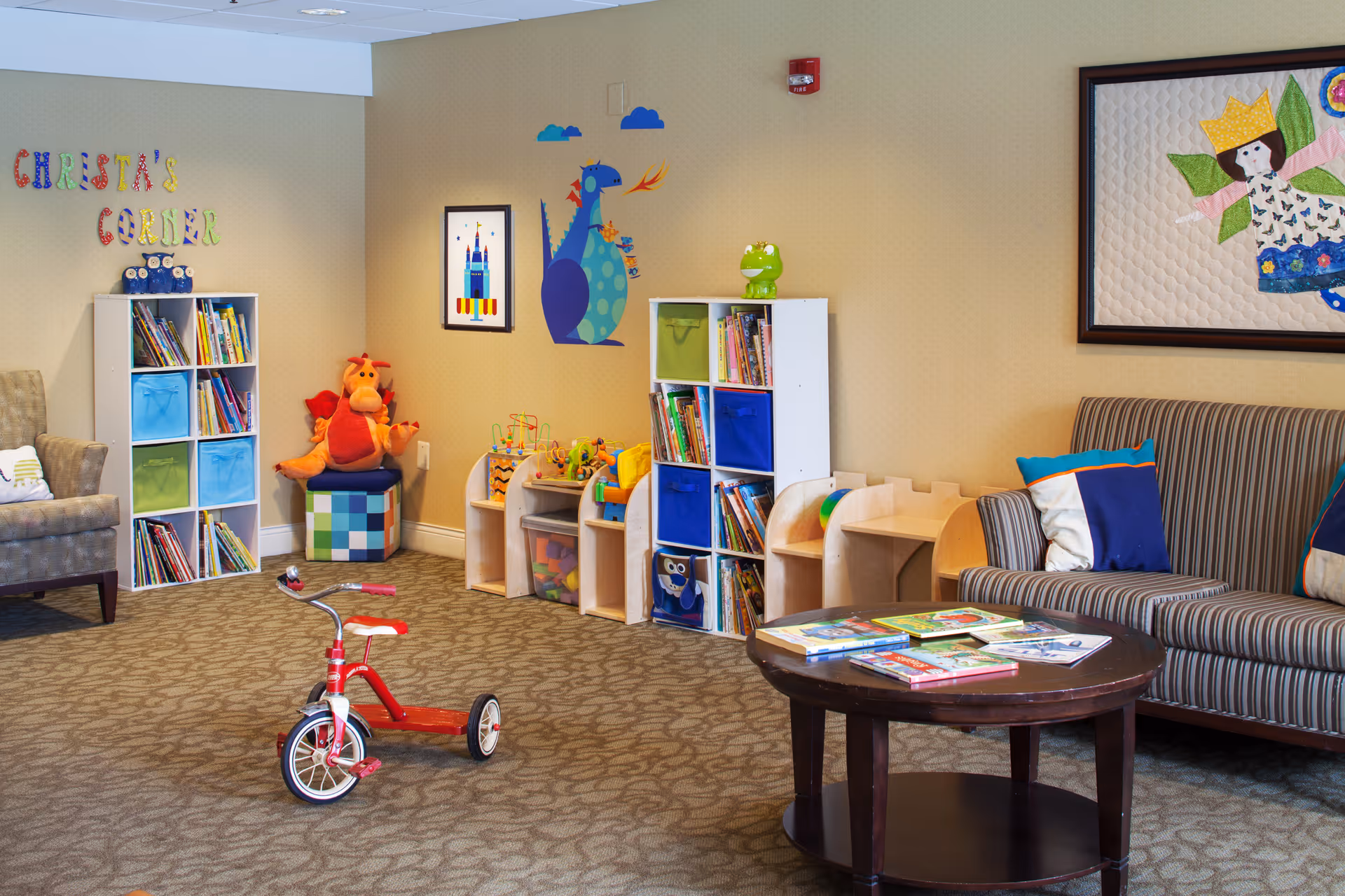 Indoor children's play corner in a common room with bookshelves, toys, a red tricycle, and seating.
