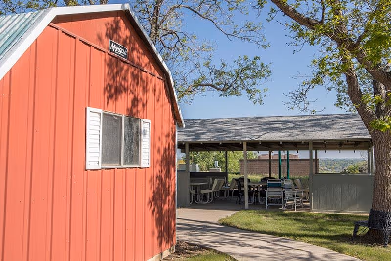 A red wooden shed with a small window and white shutters stands next to a covered outdoor seating area with tables and chairs. Trees with green leaves surround the area under a clear blue sky.