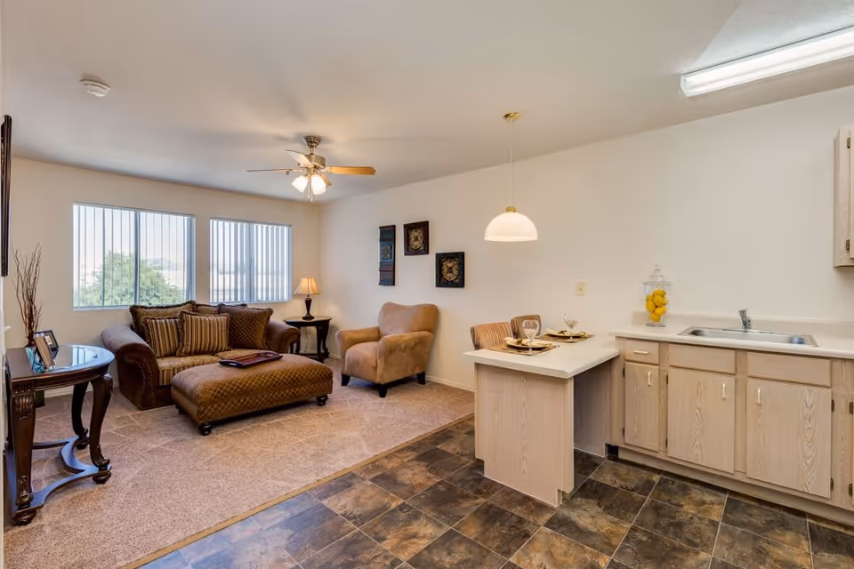 A cozy living area in a retirement community apartment featuring a brown sofa with striped cushions, a matching ottoman, a beige armchair, and a side table with a lamp. The room has two large windows with vertical blinds, a ceiling fan with lights, and a small kitchen area with light wood cabinets, a sink, and a breakfast bar with two place settings. The floor transitions from carpet in the living area to tile in the kitchen.