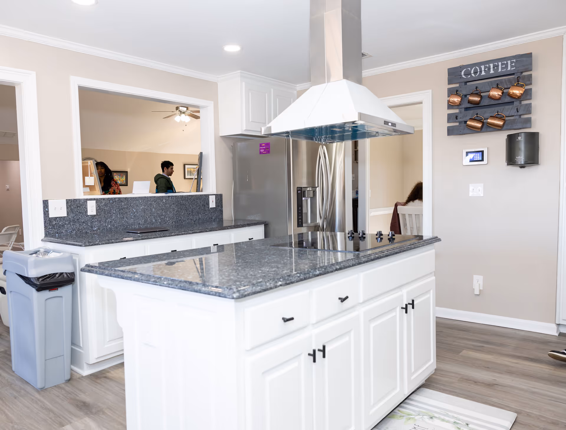 Modern kitchen area with a large island featuring a black granite countertop and a built-in stovetop. Stainless steel refrigerator is visible in the background along with white cabinetry. On the wall, there is a decorative coffee mug holder with copper mugs and the word 'COFFEE' displayed above. Two people are seen through an open window-like space connecting to another room. The floor is light wood, and a trash bin is placed near the counter.