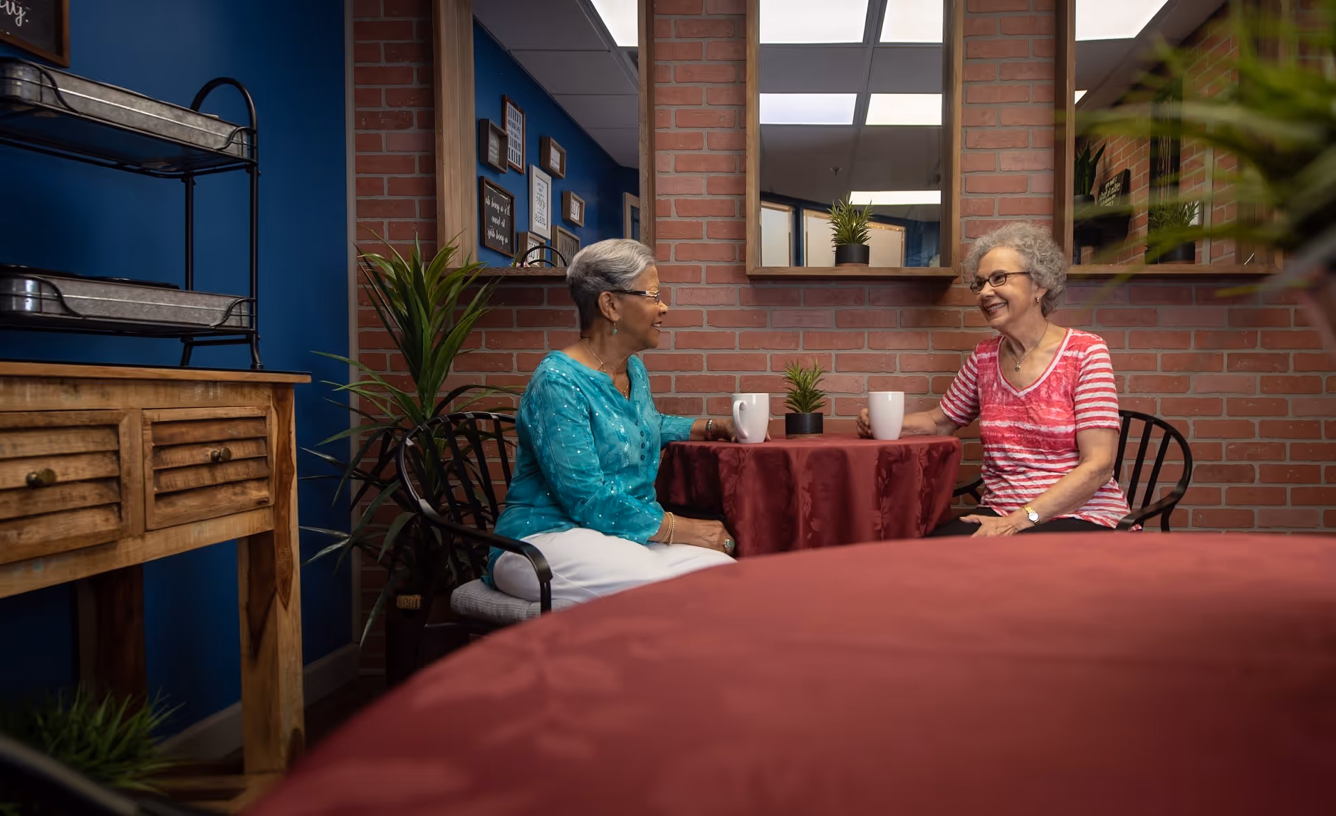 Two elderly women sitting at a small round table covered with a red tablecloth, each holding a white mug, engaged in conversation in a cozy room with brick walls, blue painted wall, wooden furniture, plants, and mirrors.