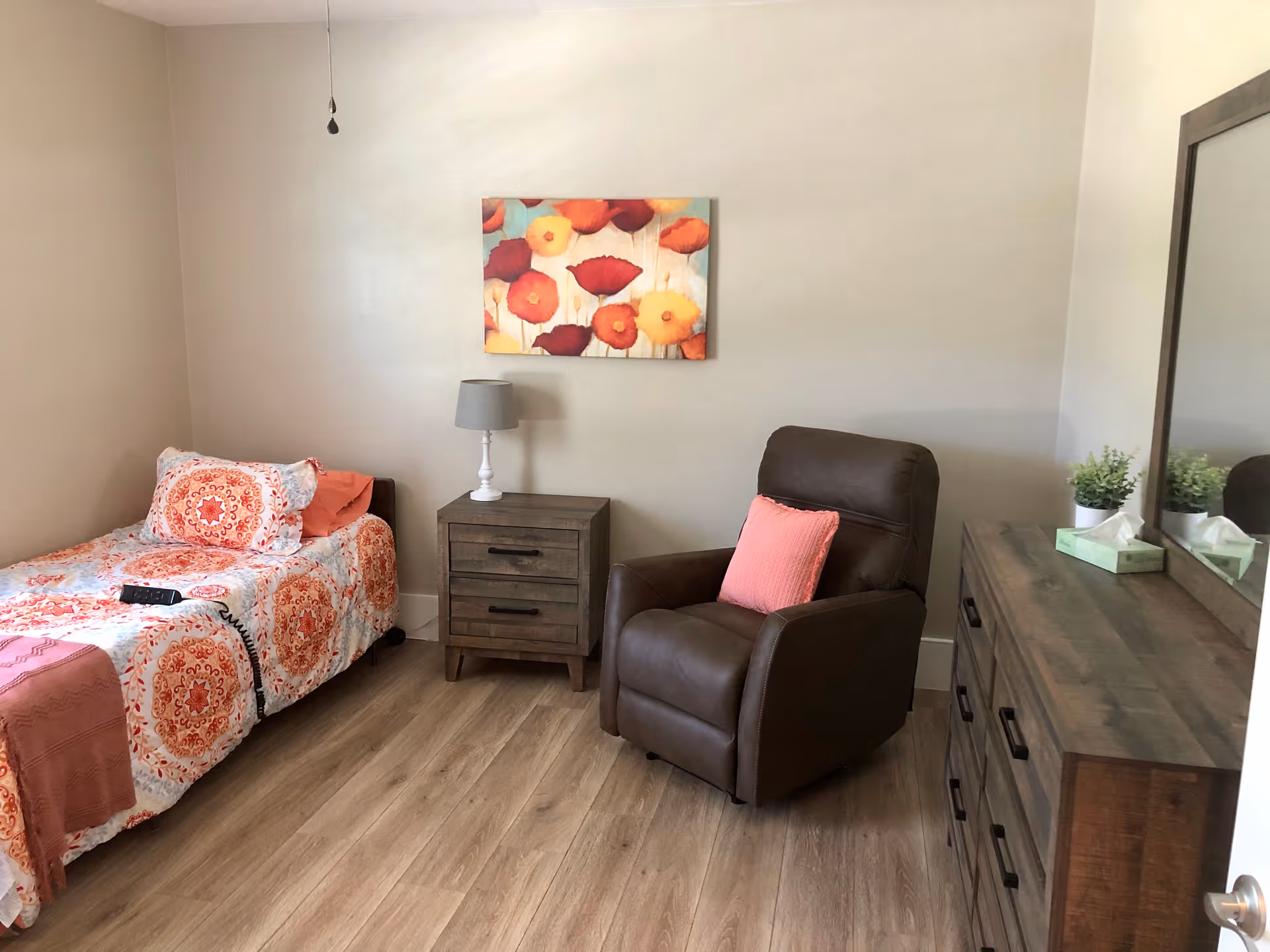 A cozy senior care bedroom with a single bed covered in a colorful orange and white patterned bedspread. Next to the bed is a wooden nightstand with a small lamp. A comfortable brown recliner chair with a coral pillow is positioned beside the nightstand. On the right side of the room is a wooden dresser with a large mirror above it, decorated with a small plant and a box of tissues. A colorful floral painting hangs on the wall above the nightstand. The room has light-colored walls and wood flooring.