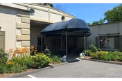 Entrance to a building with a black canopy over the doorway, surrounded by landscaped greenery and flowers under a clear blue sky.