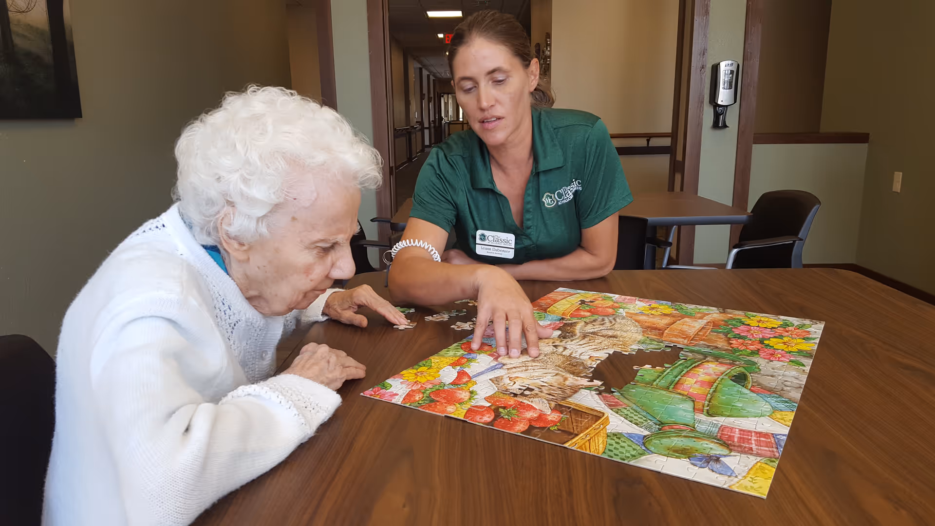 An elderly woman and a caregiver sitting at a wooden table working on a colorful jigsaw puzzle featuring a cat and flowers. The caregiver is wearing a green shirt with The Classic at Hillcrest Greens logo and a name tag. They are in a well-lit room with a hallway in the background.