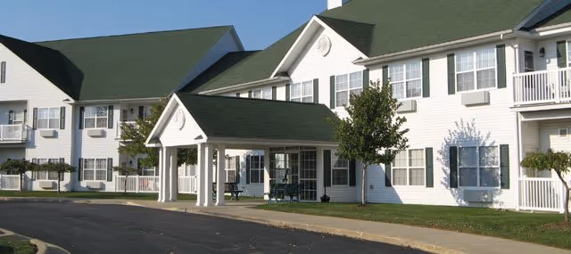 Exterior view of a white two-story senior living facility building with green roofs, multiple windows, and a covered entrance with benches underneath. There are small trees and a paved driveway in front.