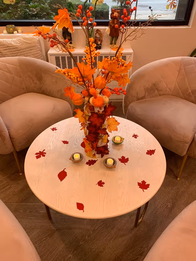 A round wooden table decorated with artificial autumn leaves and small LED candles. In the center of the table is a vase filled with fall-themed decorations including orange leaves, berries, and small pumpkins. Surrounding the table are three beige upholstered chairs. In the background, there is a window and a white radiator cover with small decorative figurines on top.