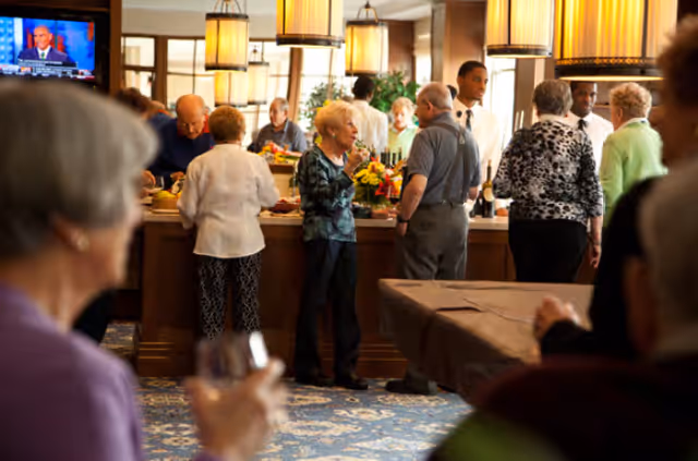 A group of elderly people socializing and standing around a buffet table in a well-lit dining area with hanging pendant lights and a patterned carpet. A television is visible in the background.