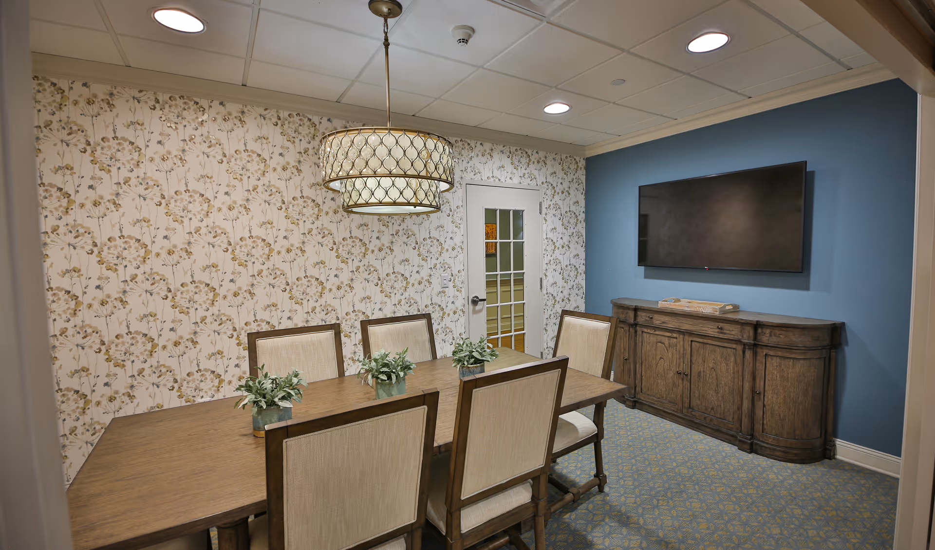 Small dining room with a wooden table and upholstered chairs, floral wallpaper, a pendant light, wall-mounted TV and wooden sideboard.
