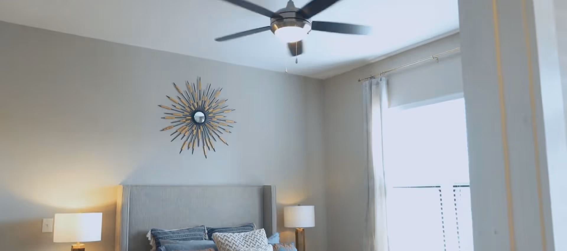 Bright, neutral bedroom interior with an upholstered headboard, decorative sunburst mirror above the bed, bedside lamps, pillows, a ceiling fan, and a window with curtains.