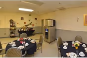 Small communal dining room with round tables set for tea, chairs, a beverage dispenser and a buffet shelf against pale yellow walls.