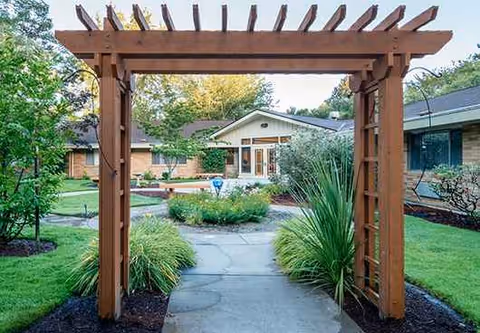 Wooden pergola frames a paved path leading to the entrance of a senior facility.
