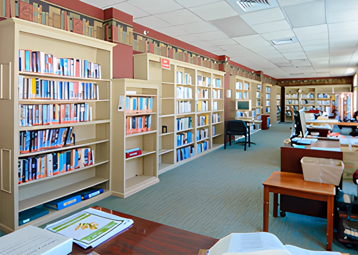 Interior view of a library or reading room with multiple bookshelves filled with books and binders along the walls. There are desks and chairs arranged in the room, with papers and books on the desks. The ceiling has white tiles and the walls are painted in a warm color with a decorative border near the ceiling.