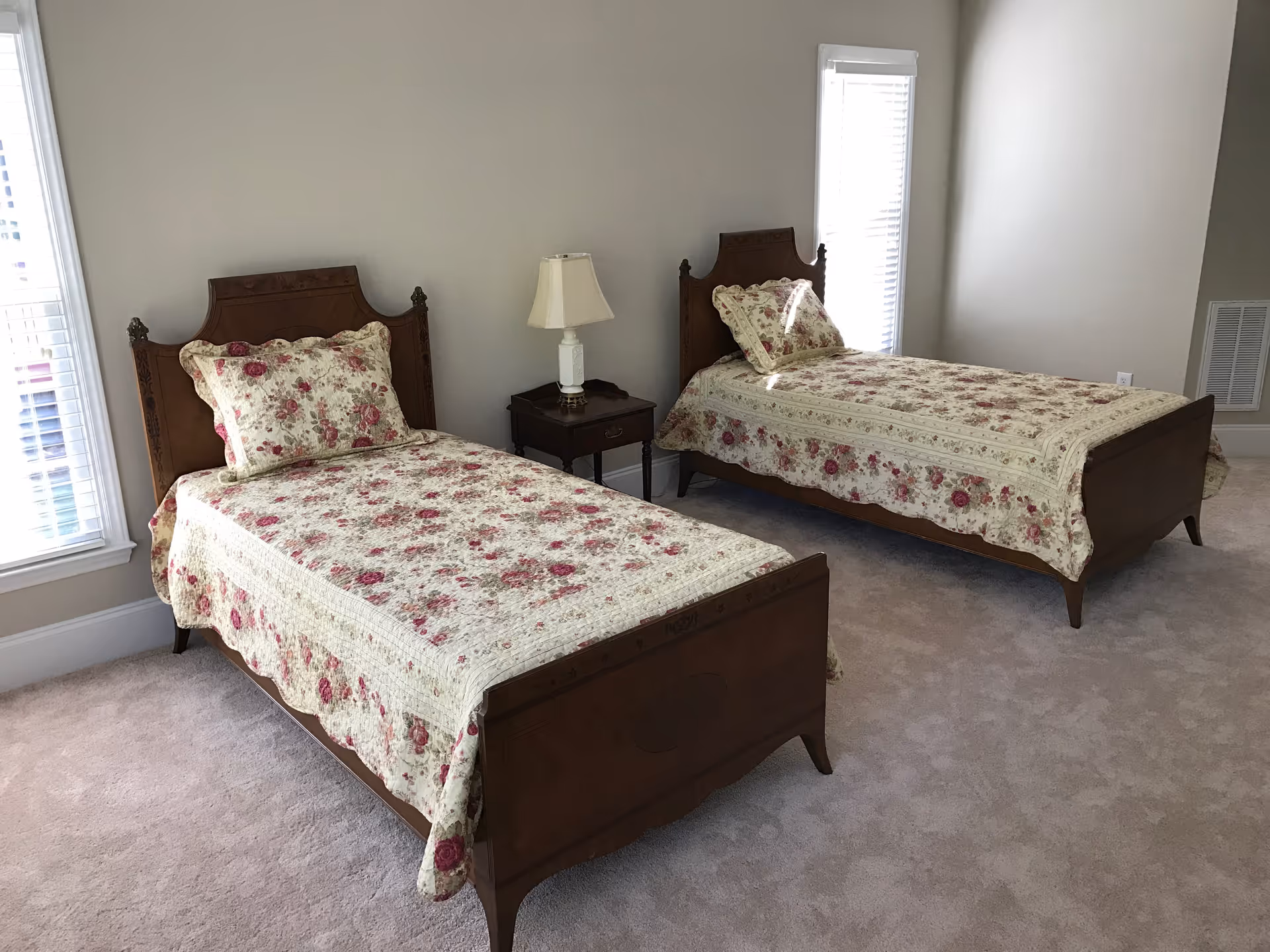 A bedroom with two twin beds featuring wooden headboards and footboards. Each bed is covered with a floral-patterned quilt and matching pillow. A small wooden nightstand with a white lamp is positioned between the beds. The room has beige walls, carpeted floor, and two windows with white blinds allowing natural light to enter.